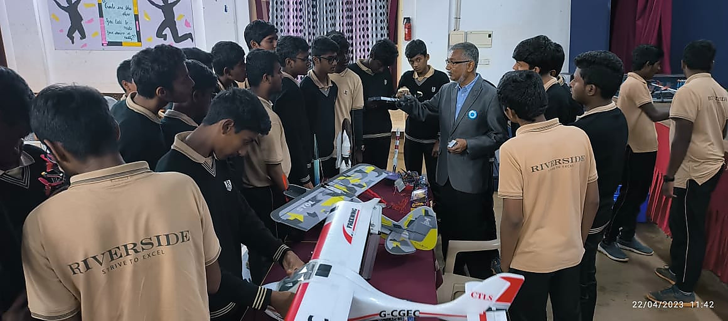 A mentor demonstrating a radio transmitter to a large circle of students around a table of RC aircraft and model rockets