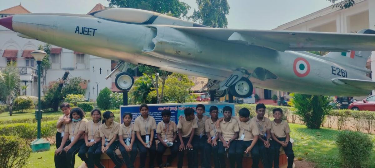 A group of school students seated beneath the preserved HAL Ajeet fighter aircraft during an aerospace visit