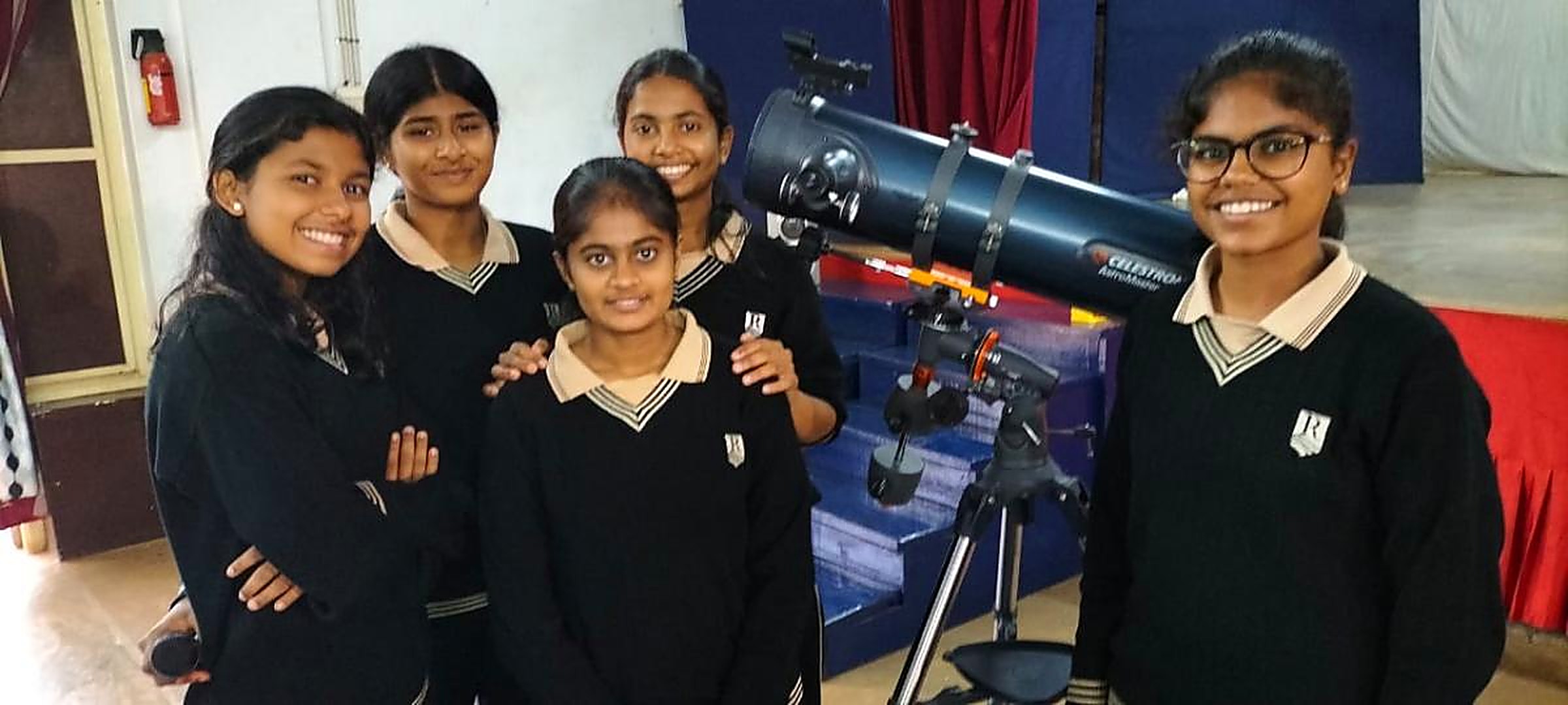 Five school students in uniform standing beside a Celestron reflector telescope set up in a school hall