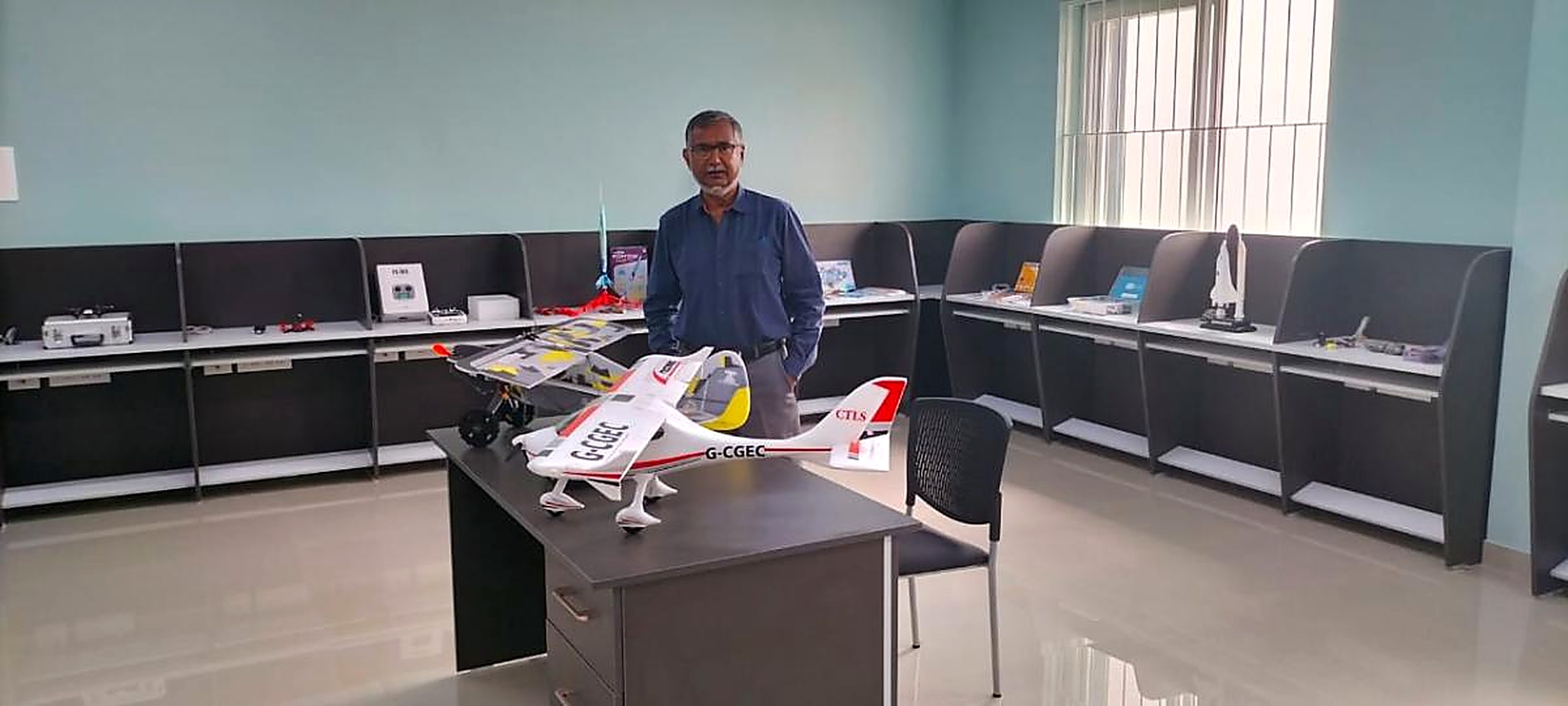A mentor standing behind a desk with a large white and red radio-controlled aircraft in an aeromodelling lab lined with display bays of kits and models