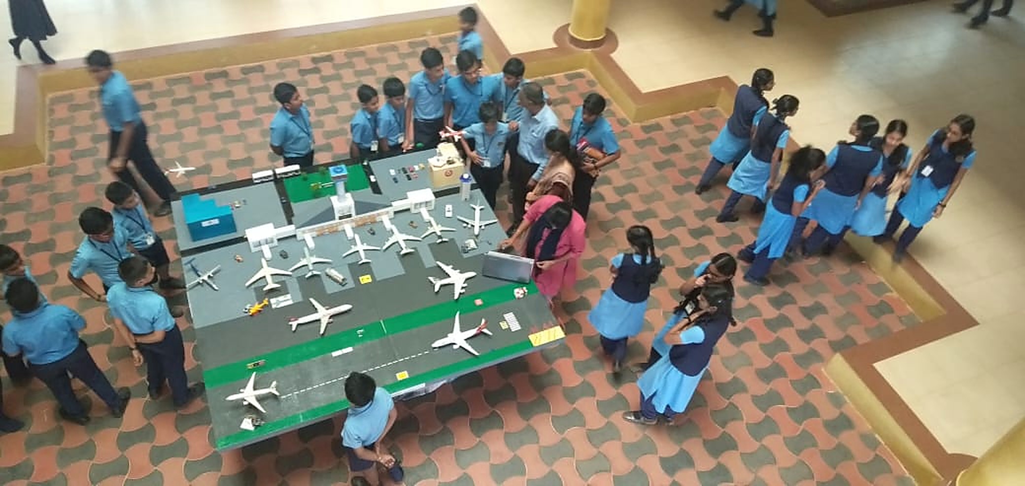 Overhead view of students in school uniform gathered around a large scale airport model with runway markings, terminals and airliner models