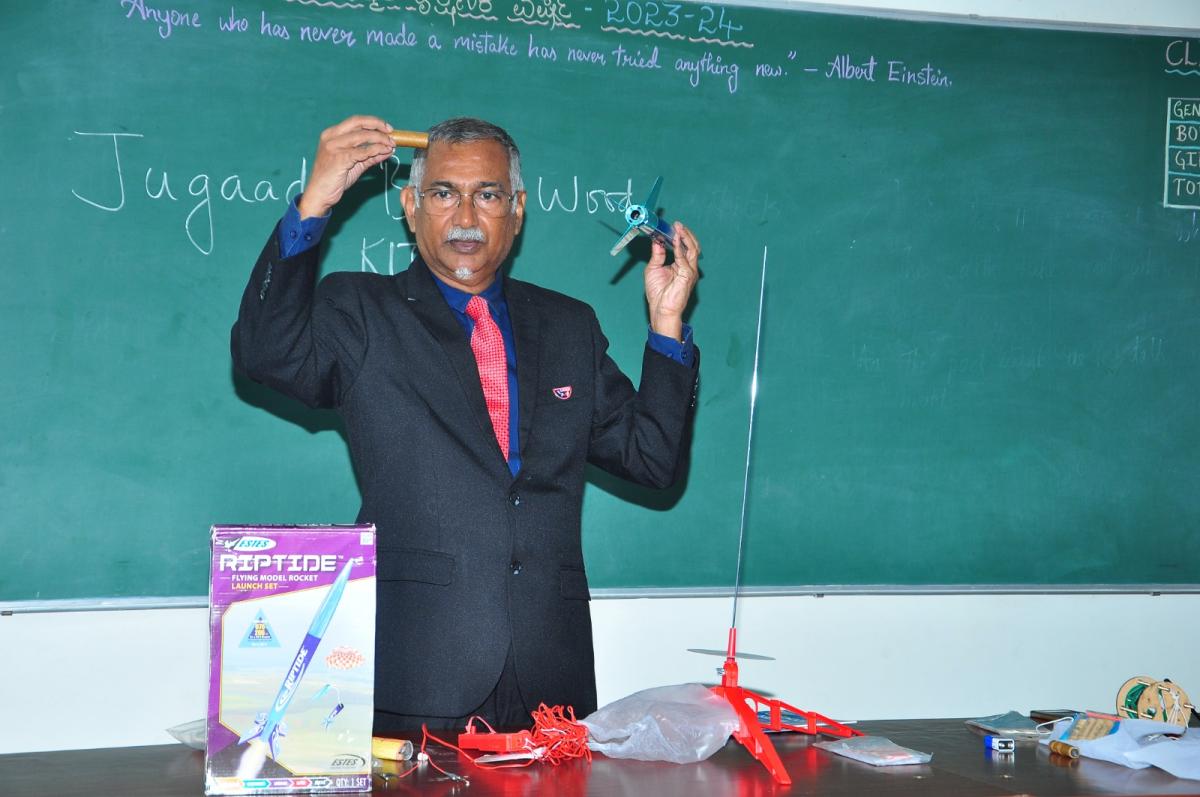 A mentor demonstrating a model rocket motor and component beside a launch kit and pad in front of a classroom blackboard