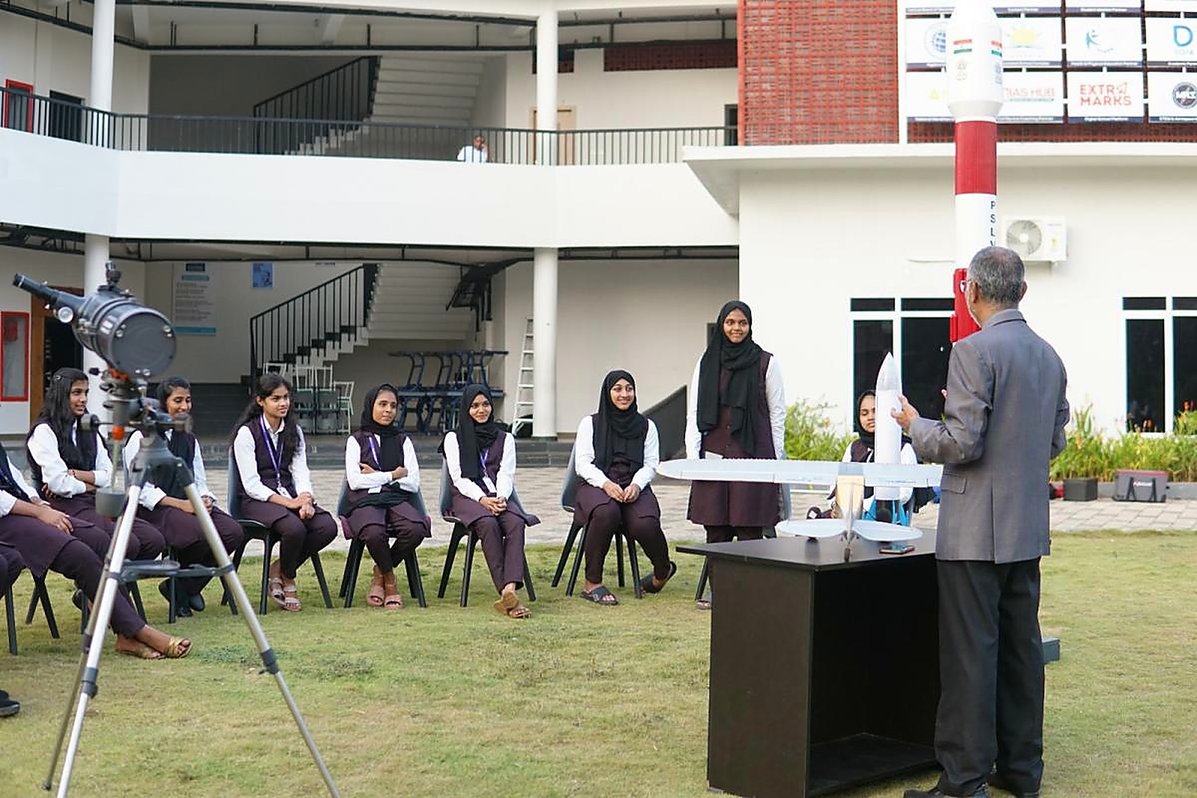 A mentor holding a rocket model in front of seated students on a lawn, with a telescope on a tripod and a full-size PSLV model behind
