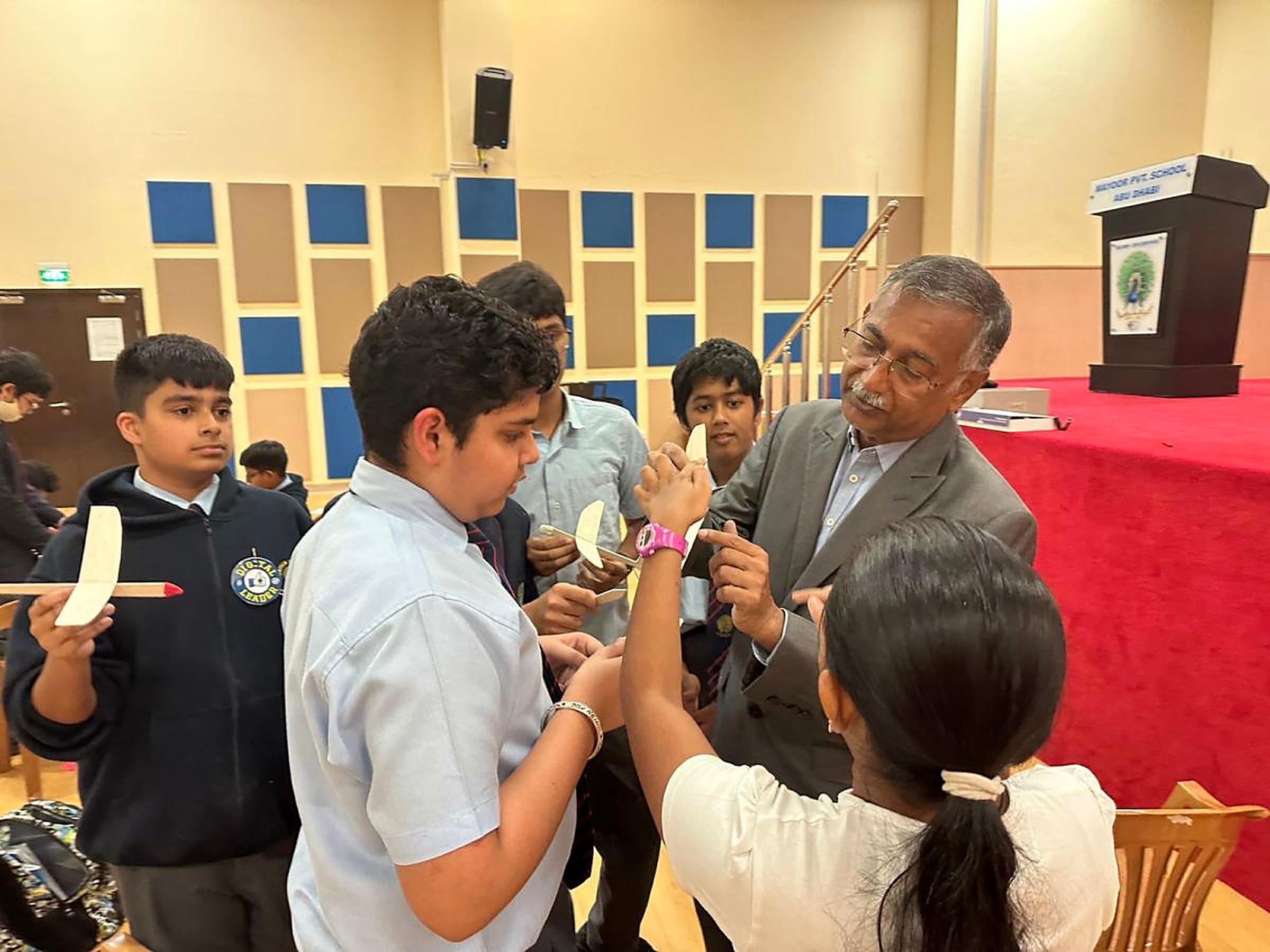A mentor guiding a group of school students as they hold and adjust balsa-wood glider models during an MRS aeromodelling session in a school hall