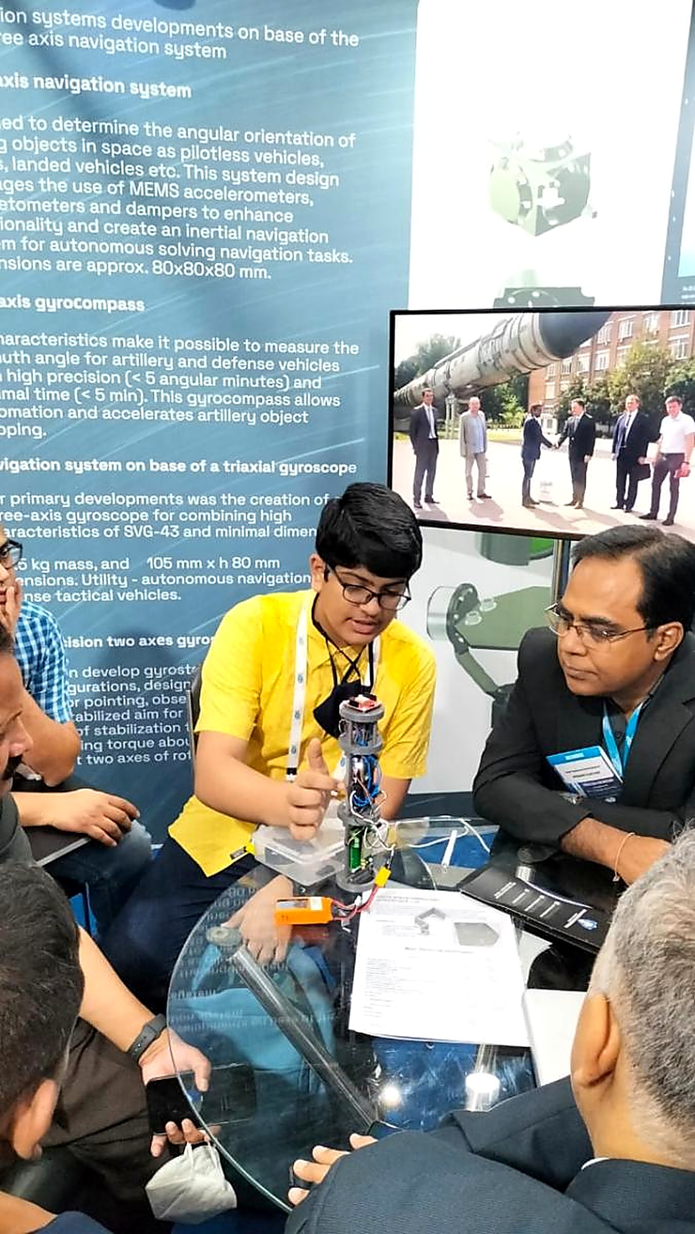 A student explaining a three-axis gyroscope payload module to industry delegates seated around a table at a space expo stand