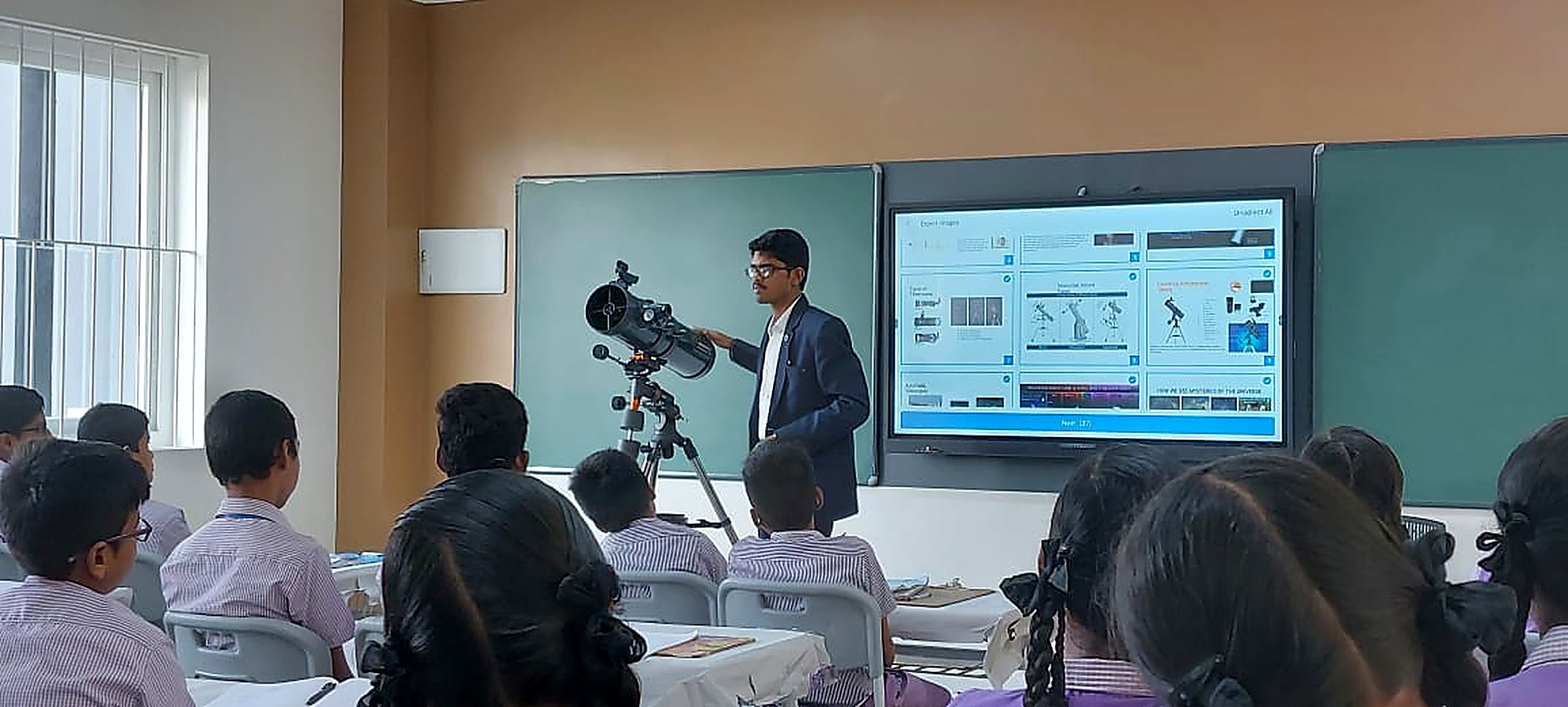 A young mentor in a blazer presenting a reflector telescope beside a large screen of astronomy slides to a classroom of primary school students