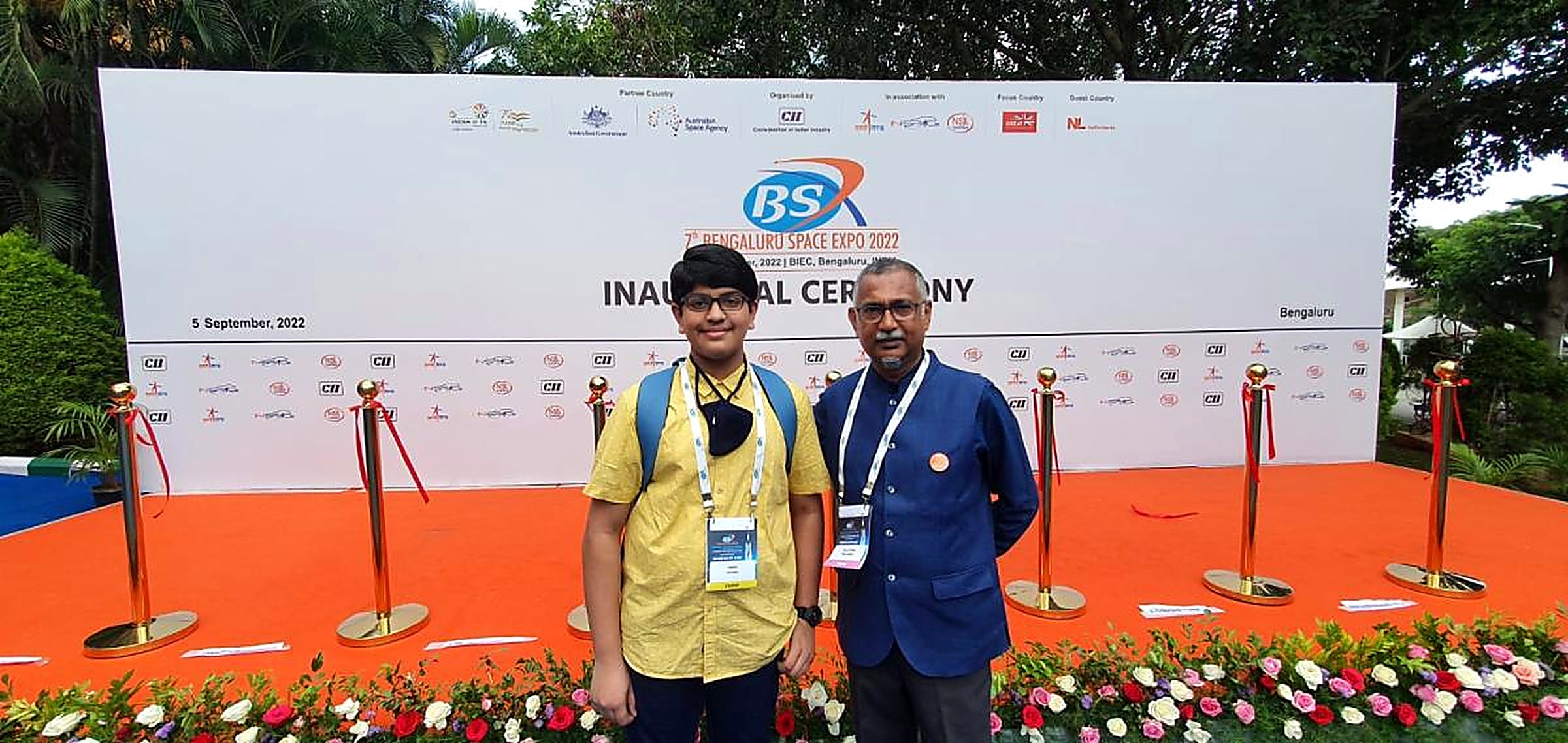 A student and a mentor standing in front of the inaugural ceremony backdrop of the 7th Bengaluru Space Expo 2022