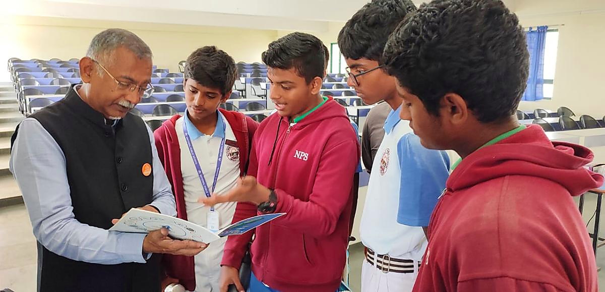 A senior mentor showing a technical booklet to five attentive students in an empty lecture hall