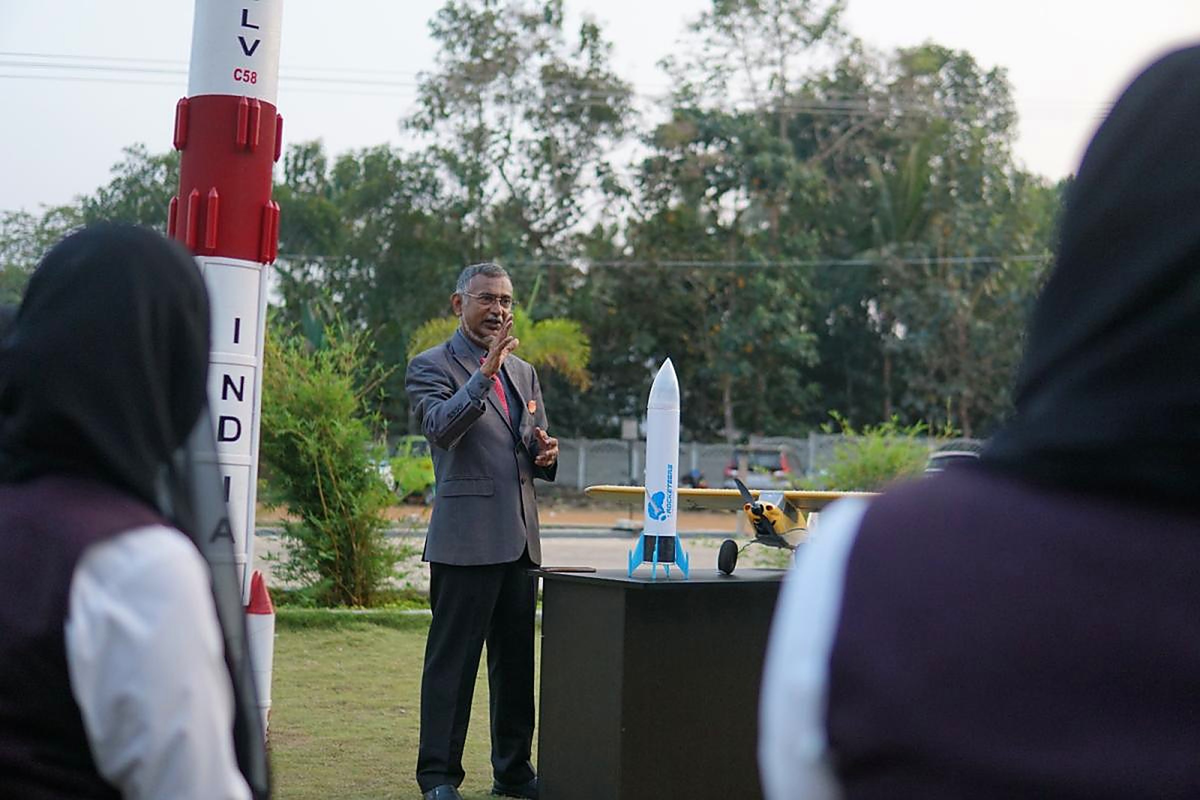 A mentor explaining rocket staging to students on a lawn, standing between a full-size PSLV C58 model and a table with a model rocket and RC aircraft