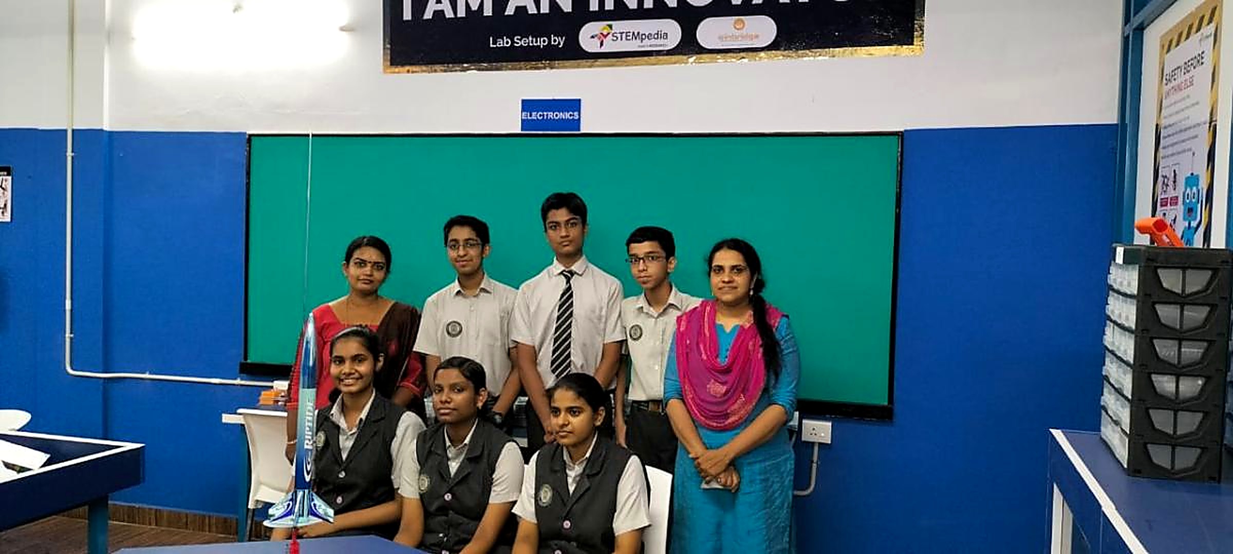 A school team of eight students and two teachers in an innovation lab with a blue and white model rocket on the table in front of them