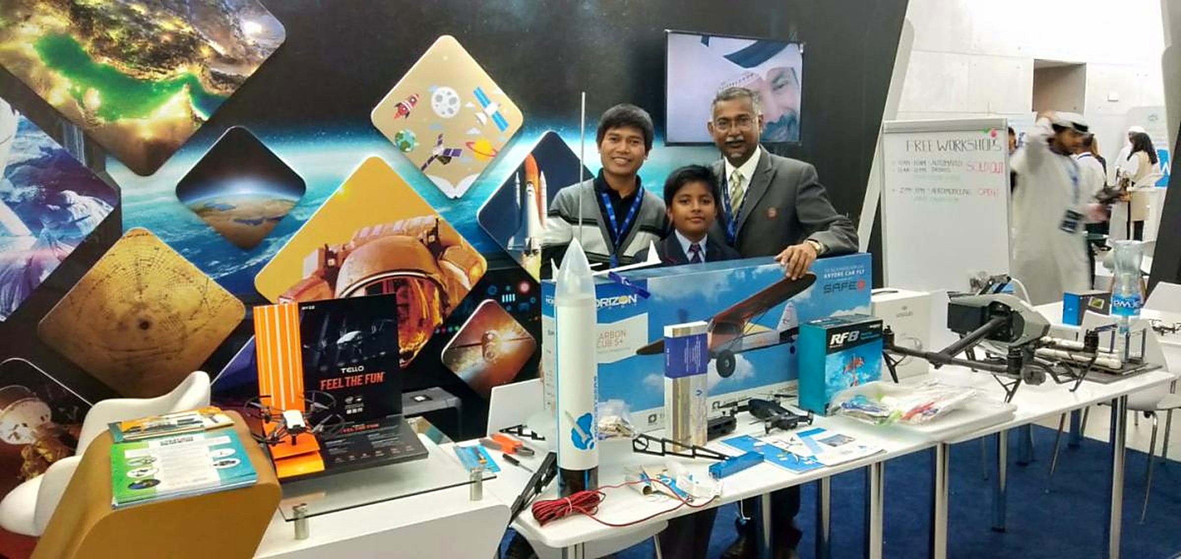 A mentor, an instructor and a young student at an MRS aerospace stand displaying a model rocket, RC aircraft kits and drones