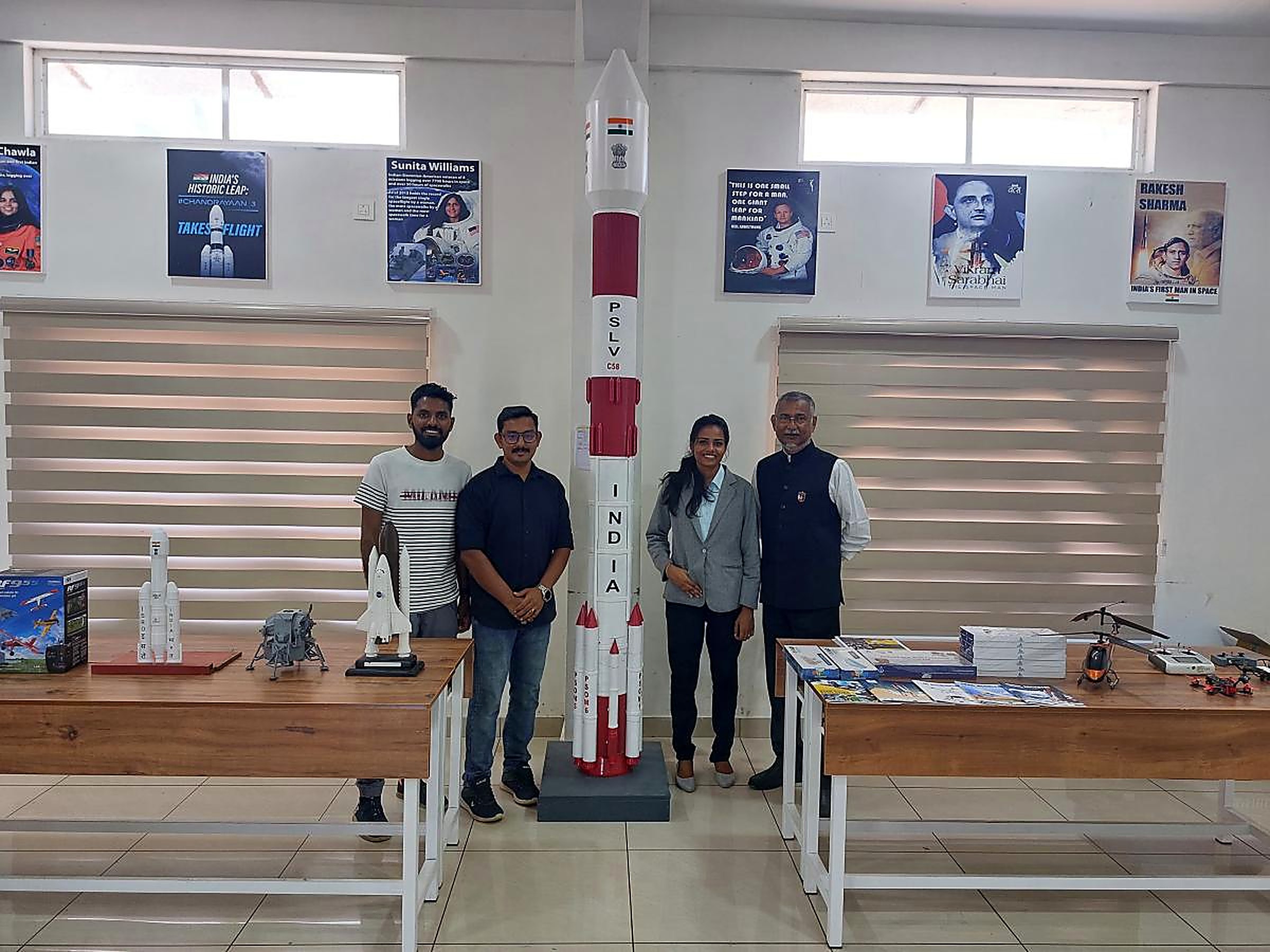 Four MRS team members standing beside a tall PSLV C58 model in a space lab lined with astronaut posters and tables of rocket, lander and drone models