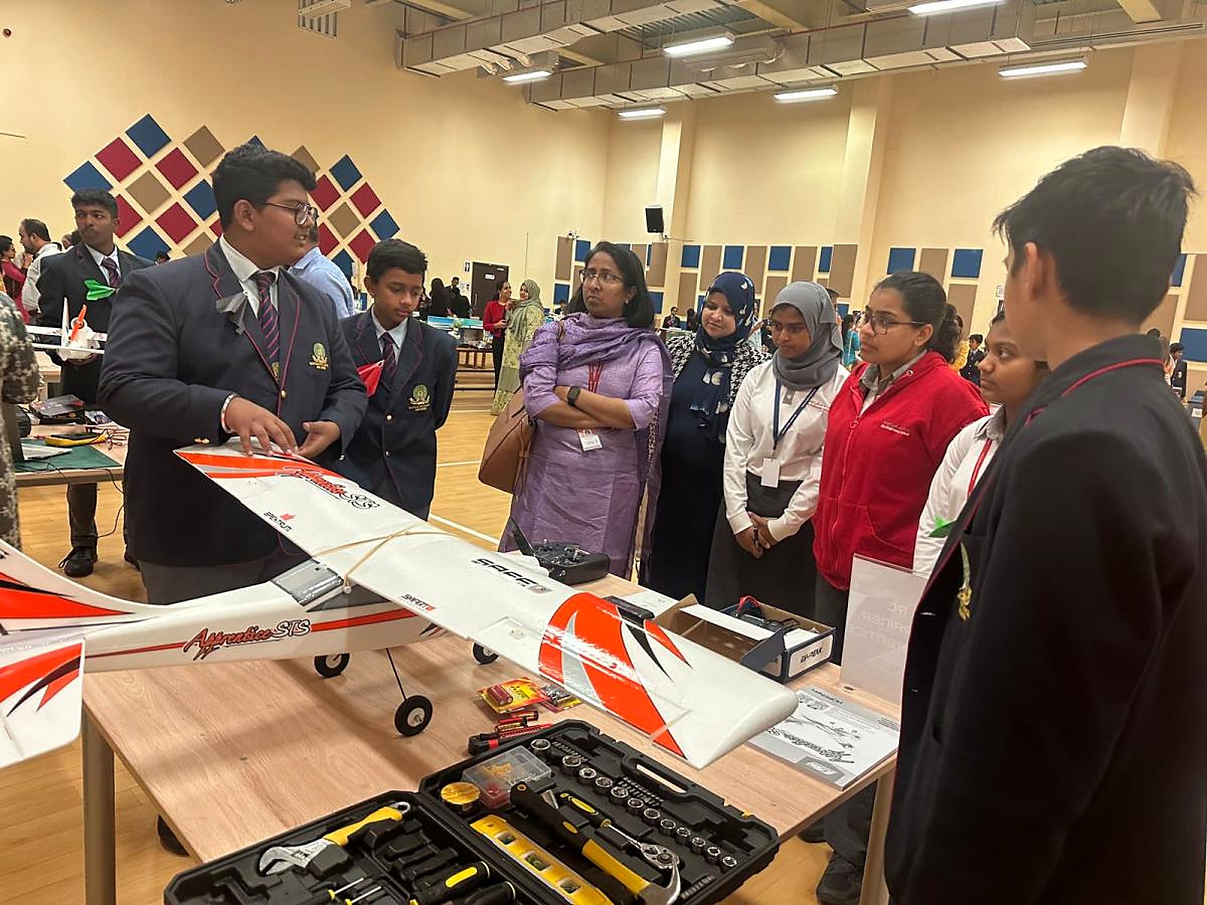 A student in a blazer presenting a red and white radio-controlled aircraft on a table to teachers and fellow students at an exhibition
