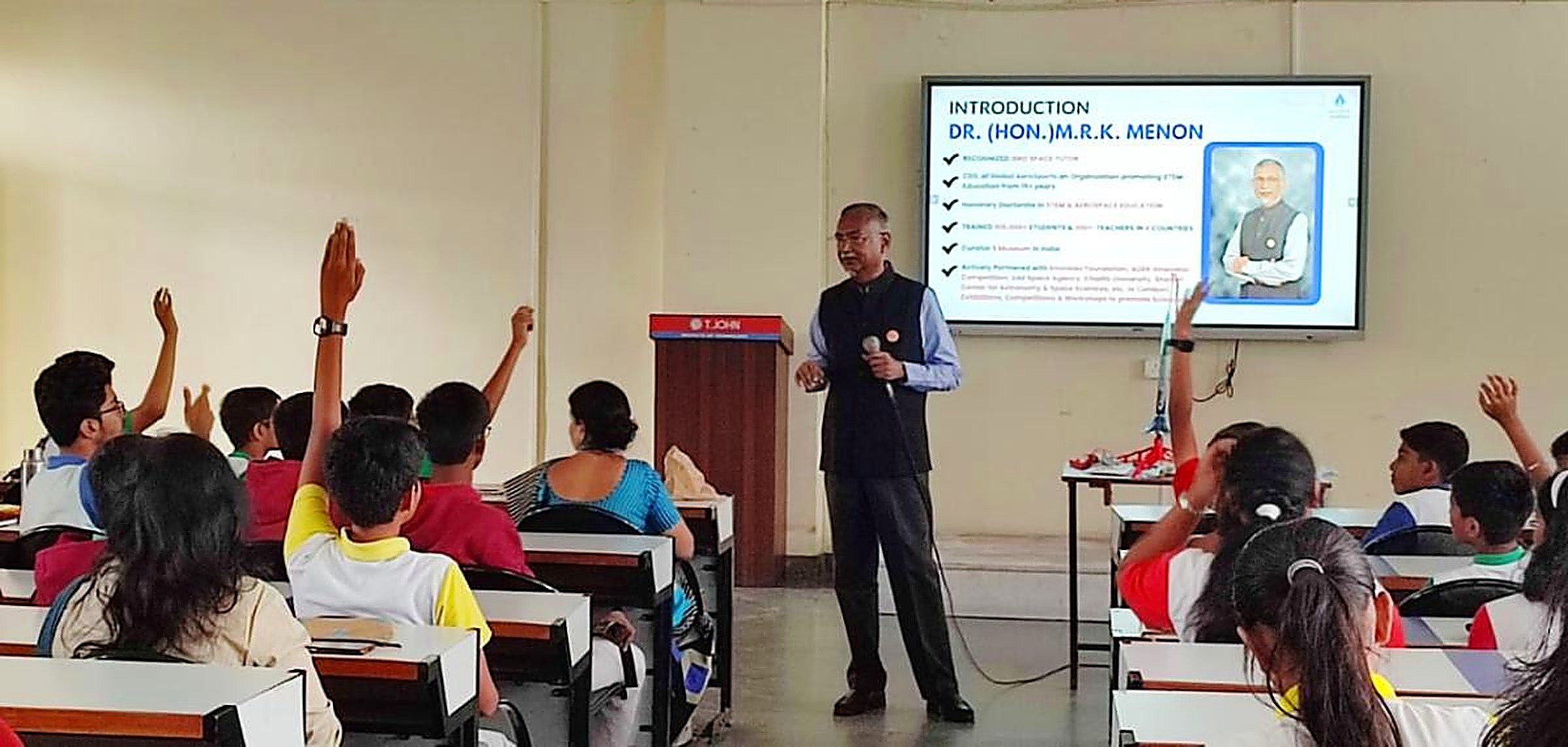 Dr. M. R. K. Menon addressing a lecture hall of school students who are raising their hands, with his introduction slide on the screen behind