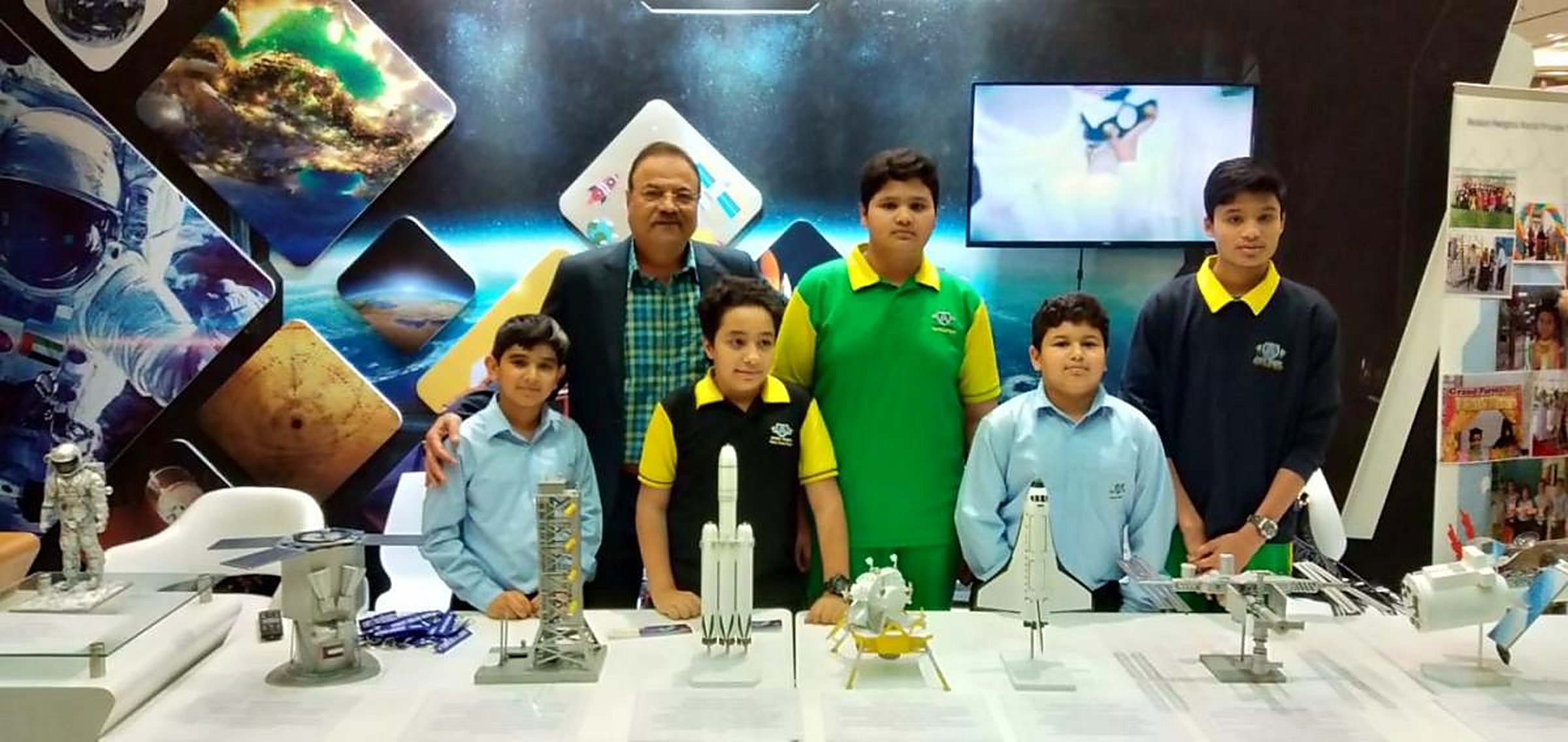 A mentor standing with five students behind a display table of launch vehicle, lander and space shuttle models at a space exhibition stand